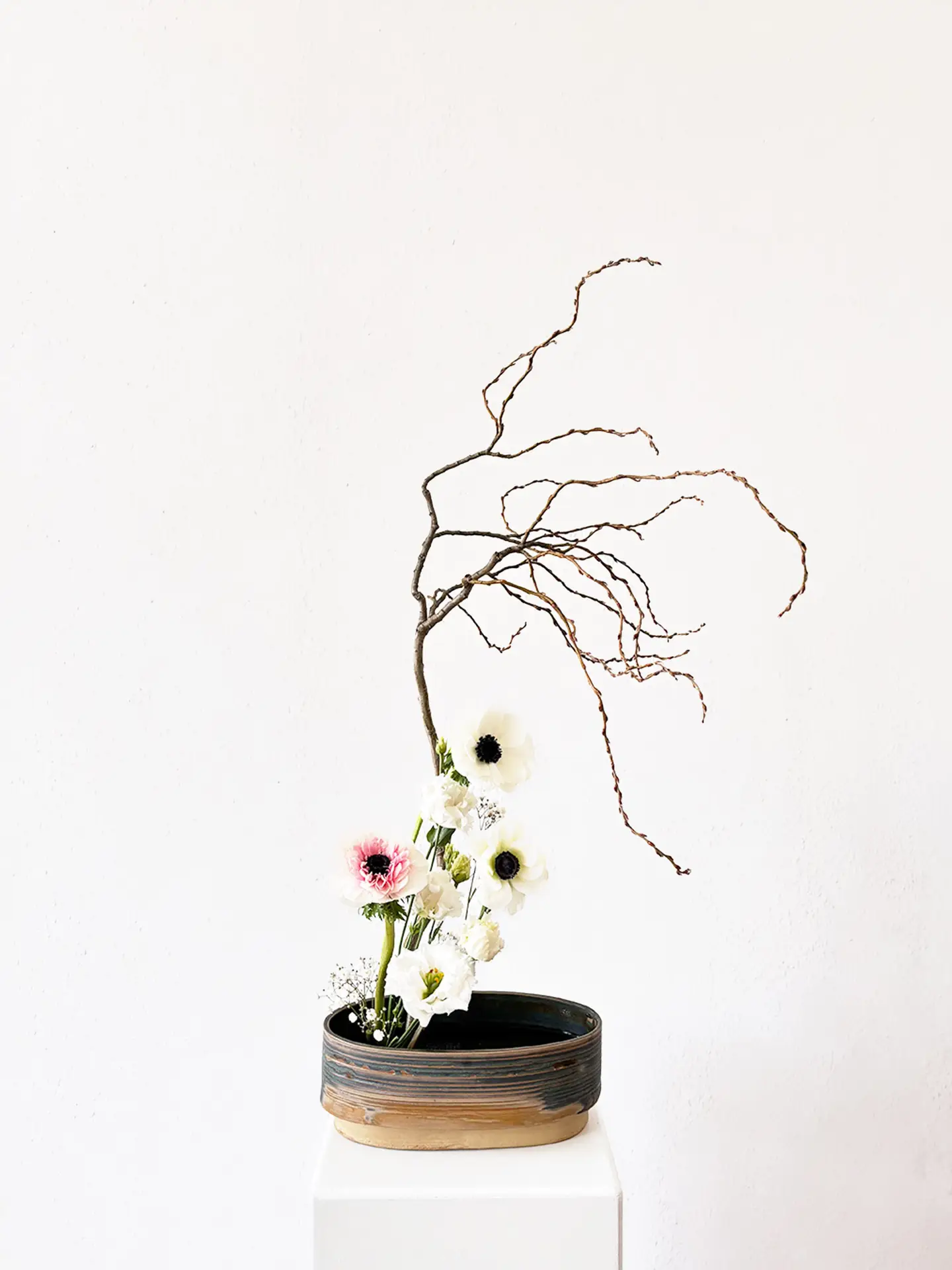 Sculptural ikebana arrangement with white anemones and a single pink accent bloom, paired with a dynamic bare branch in a handmade ceramic vase by Verena De Hoog, created by suminumi in Berlin.